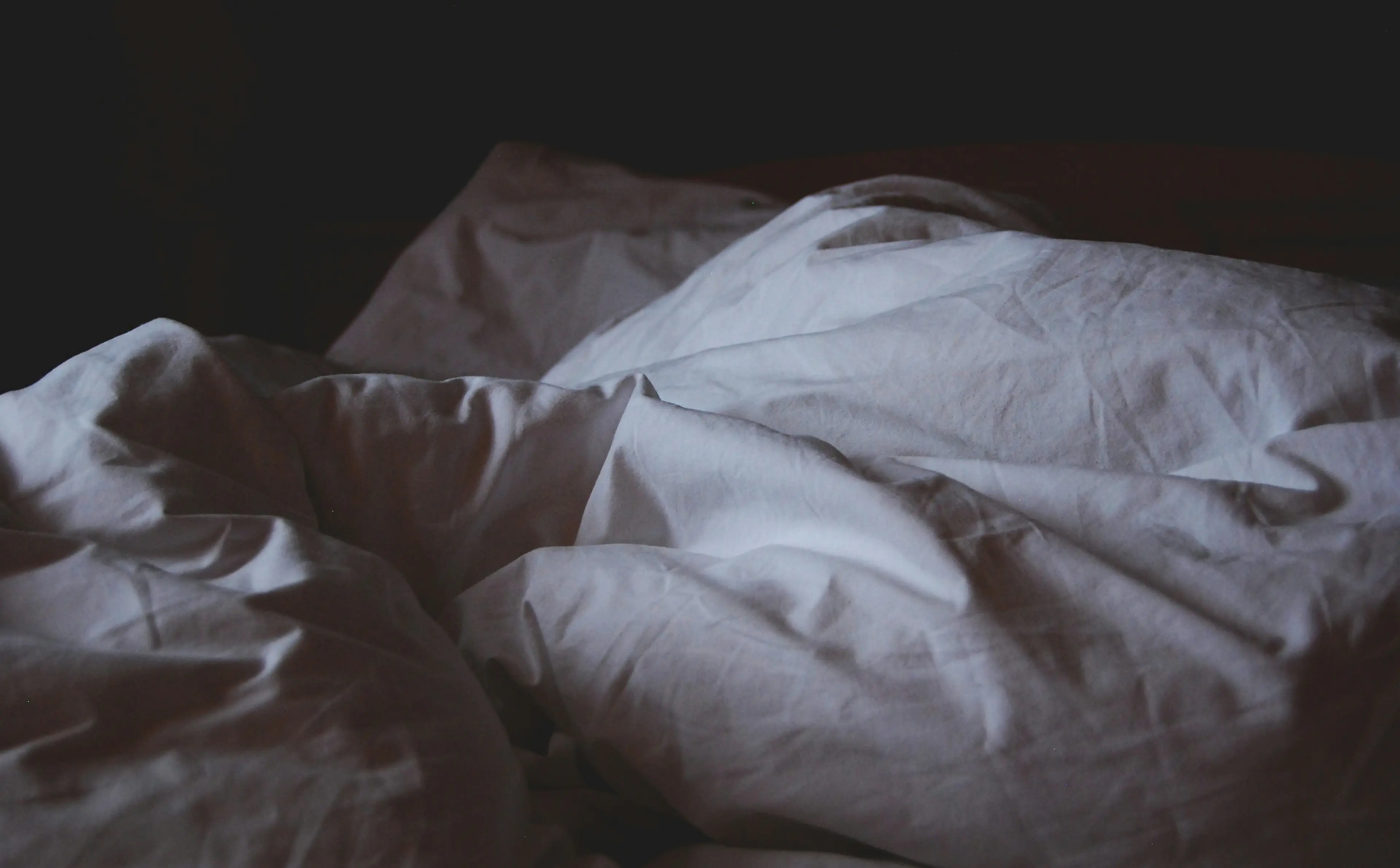 Leeres Bett mit zerwühlter weißer Decke und Kissen in einem abgedunkelten Schlafzimmer - Hypnose zum Einschlafen